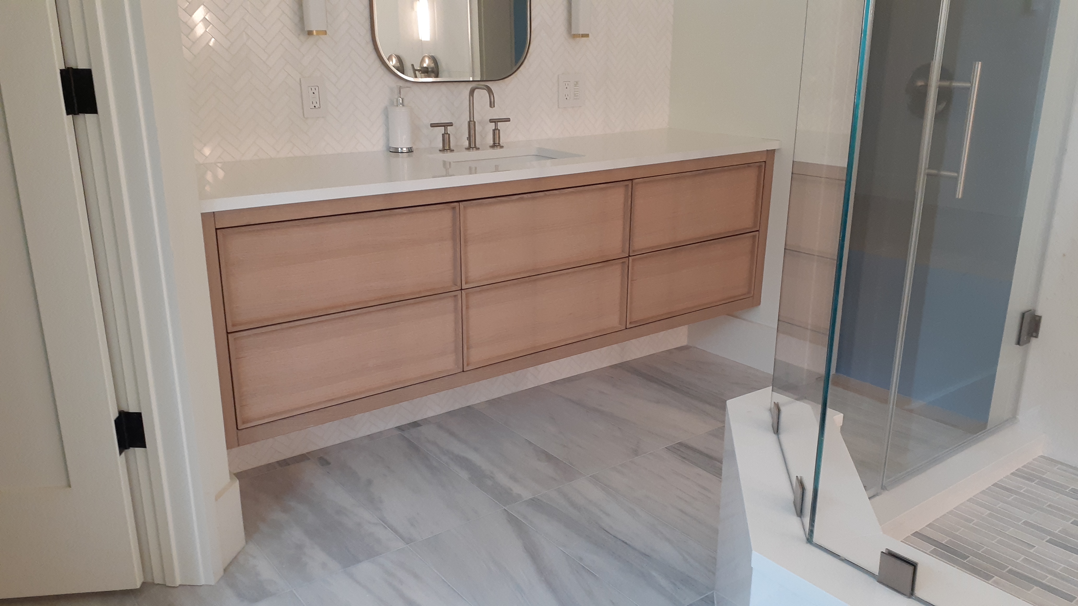 Floating natural wood bathroom vanity with six drawers, white countertop, herringbone tile wall, and glass shower beside it
