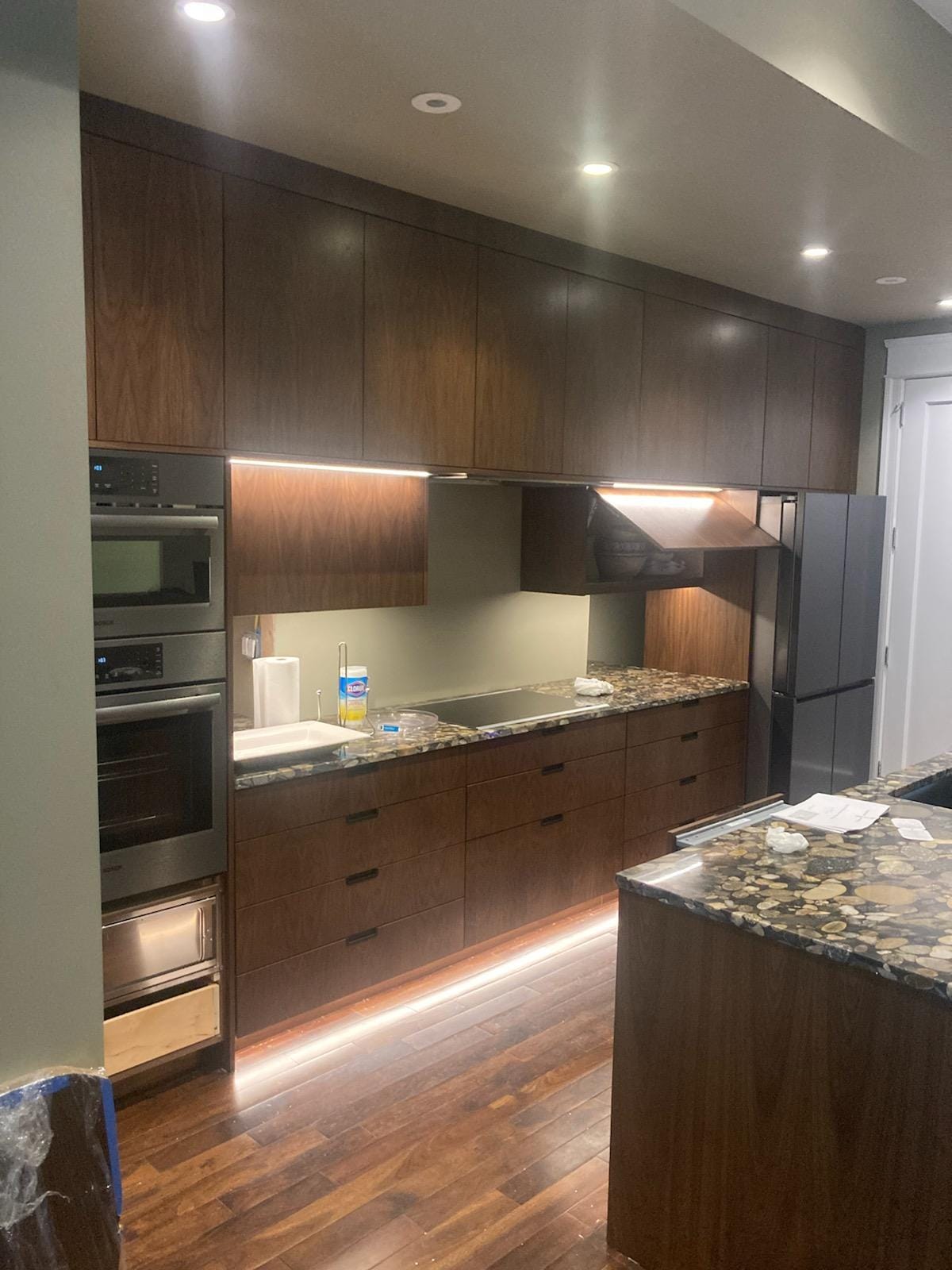 Contemporary dark wood kitchen with full-height cabinets, stacked ovens, and illuminated work areas