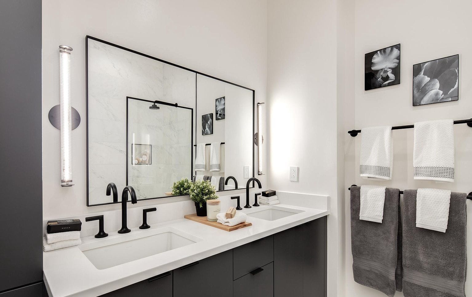 Modern double bathroom vanity with dark cabinet fronts, white counter, black faucets, large mirror, and vertical sconces