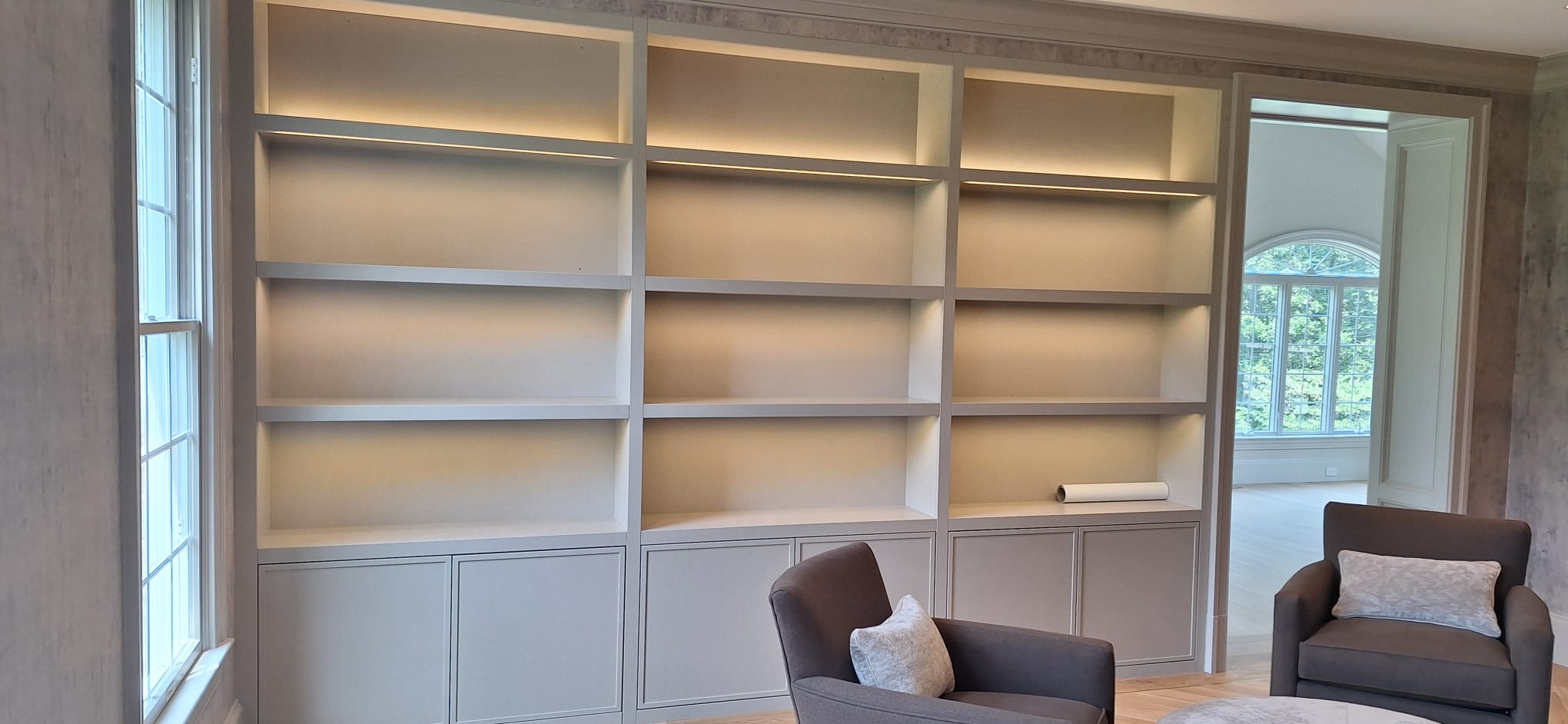Light gray floor-to-ceiling built-in shelving with lower cabinet doors and integrated shelf lighting in a sitting room