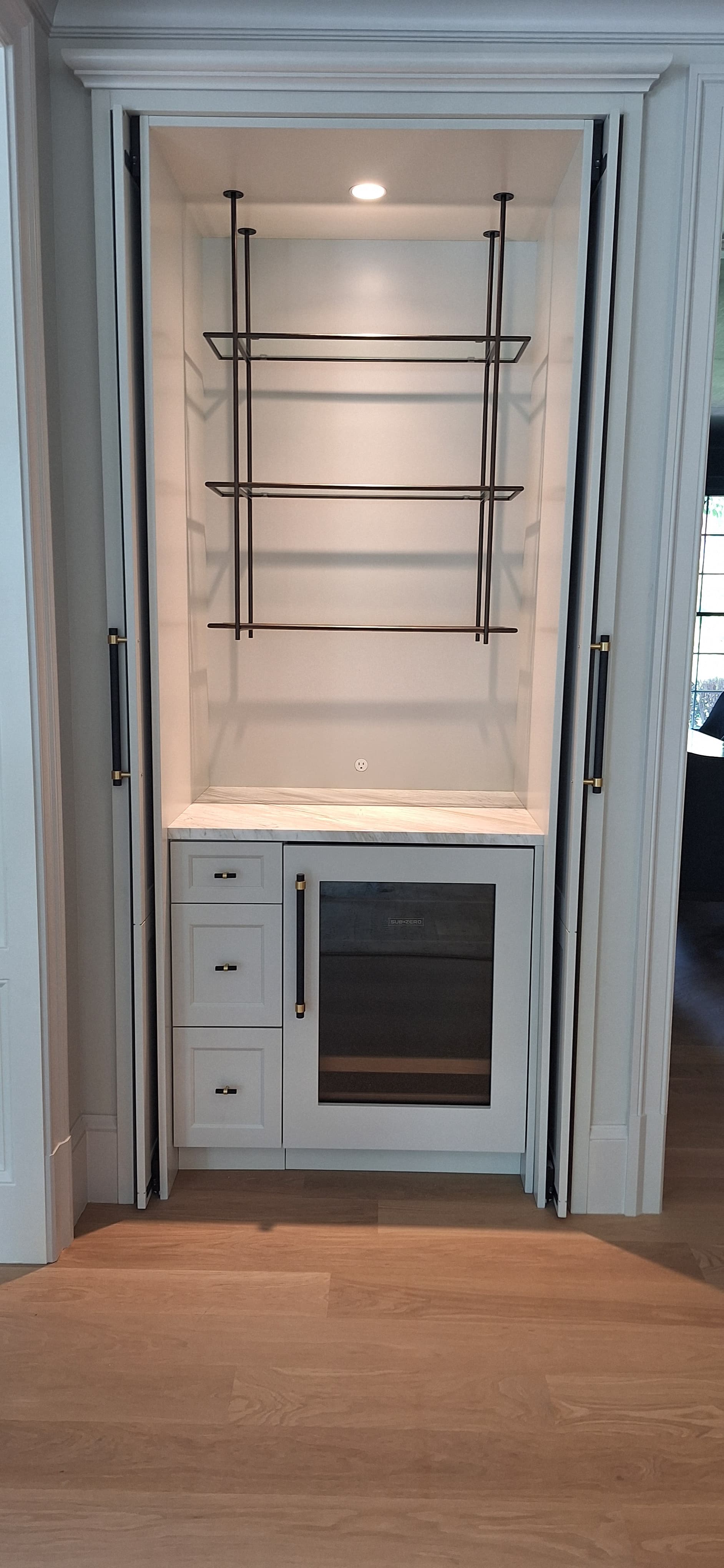White bi-fold cabinet doors open to reveal a hanging stemware rack, marble counter, and built-in wine refrigerator