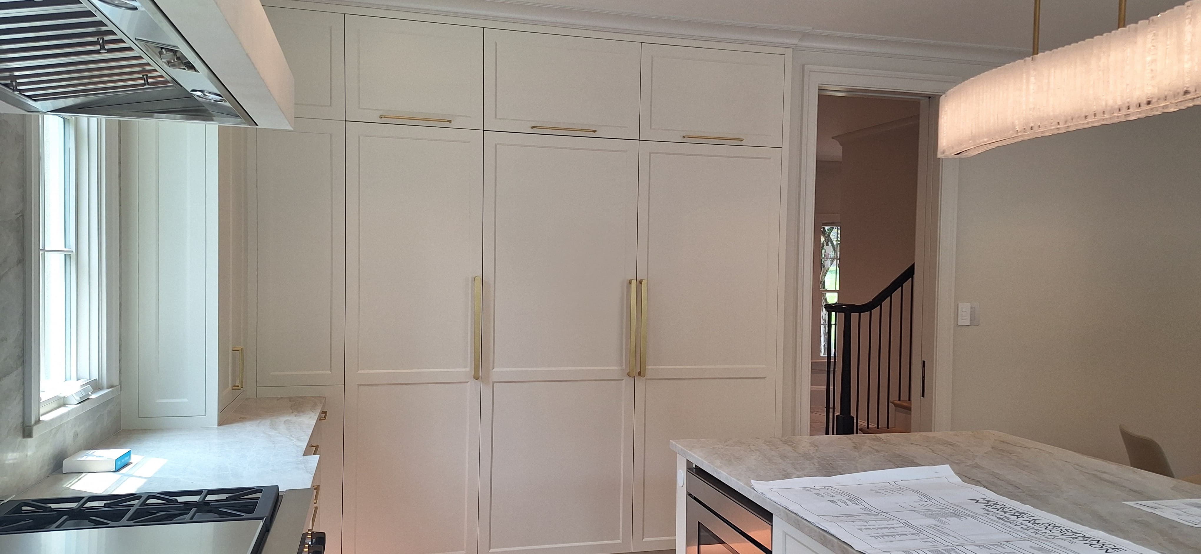 Tall pantry cabinets with brass pulls beside a range hood, opening toward a staircase hallway
