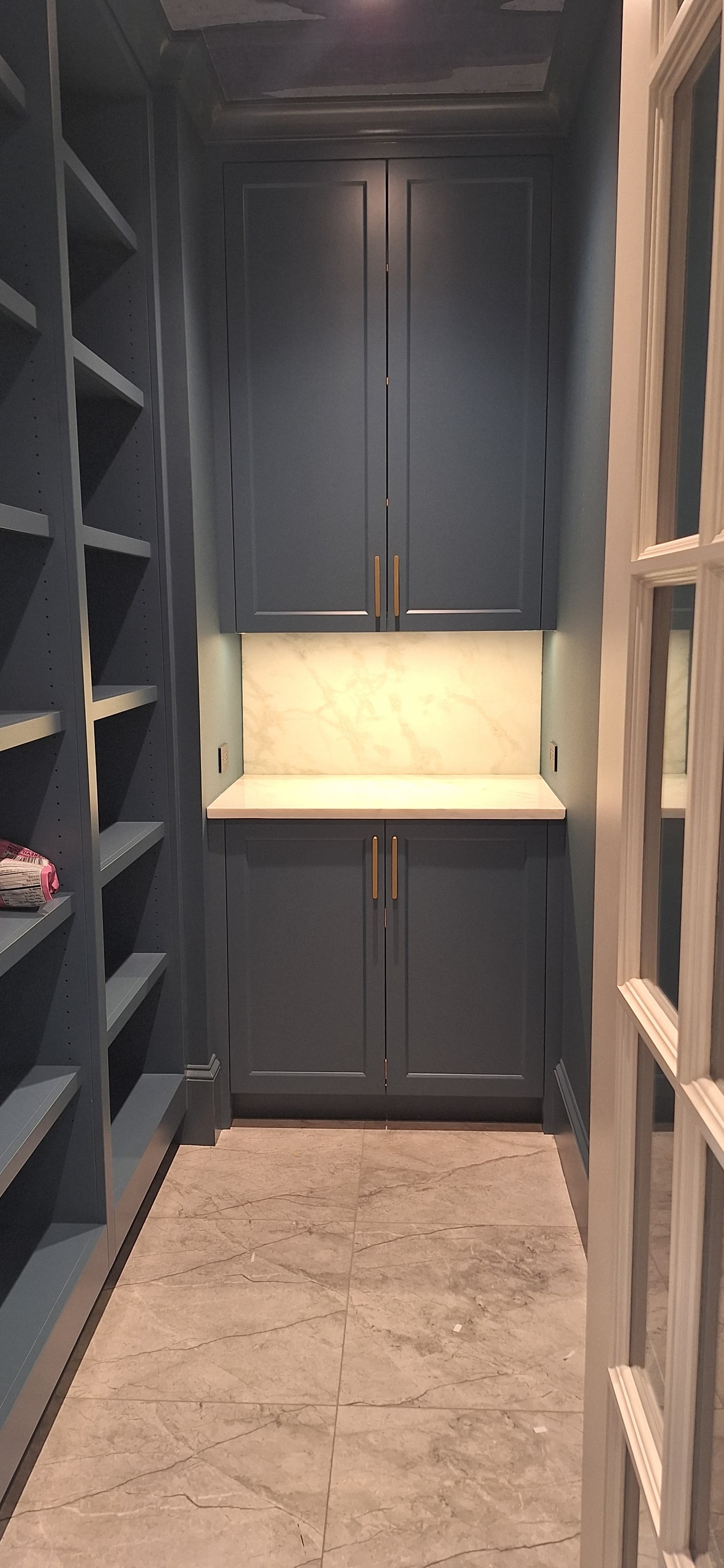 Dark blue-gray pantry cabinetry with a marble backsplash and hanging stemware rack at the end of a walk-in pantry